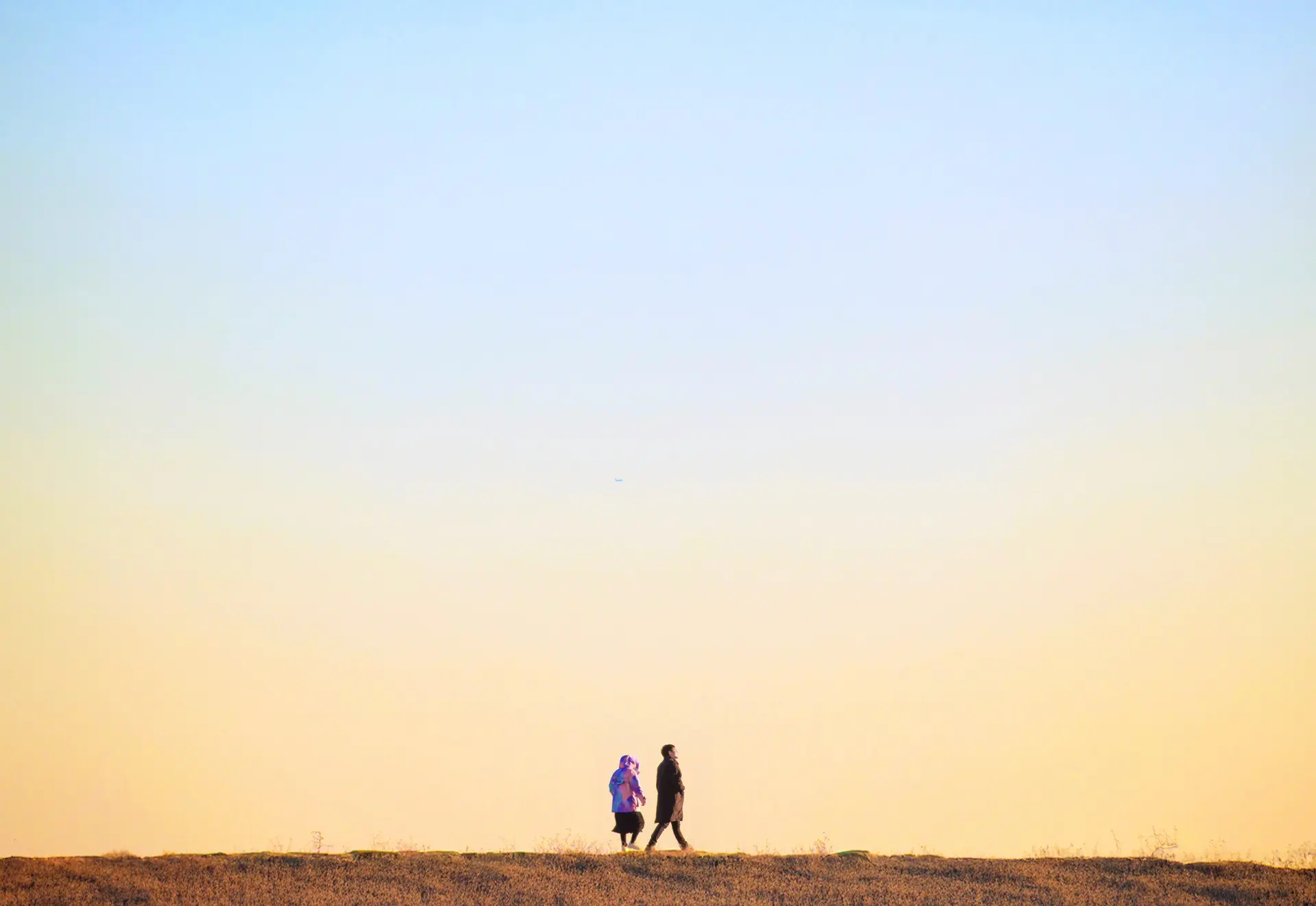 Silhouettes of two people stand on a flat landscape against a gradient sky at dusk.